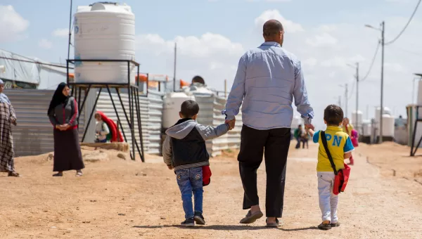 A father walks down the road of the camp he lives in with his family whilst holding his children's hands.