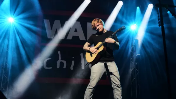 Ed Sheeran standing on a stage playing his guitar with the black, red and white War Child logo in the background. Simple white spotlights dot through the stage