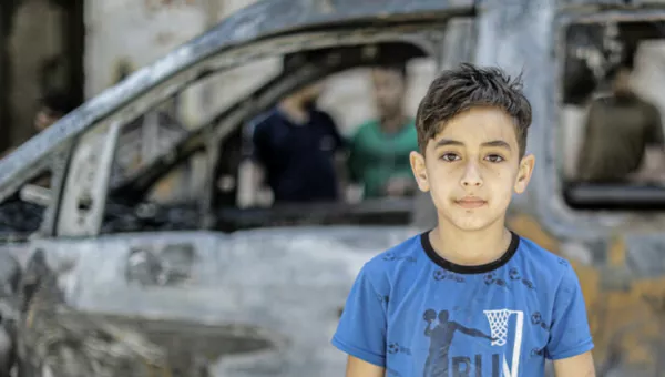 A young boy in a blue t-shirt standing in front of a car in burnt out car