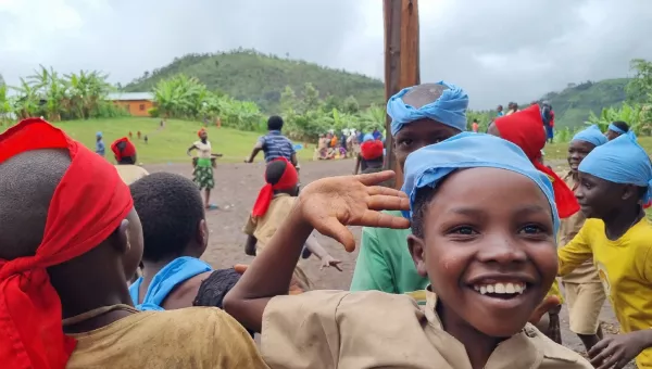 Two teams of children playing a game of tag in Burundi with red and blue bandanas on their heads. A child looks to the camera smiling with their hand raised in the air.