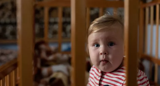 Ukrainian baby sits in cot looking at camera