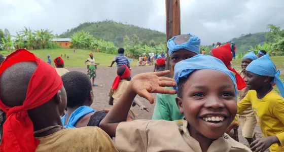 Two teams of children playing a game of tag in Burundi with red and blue bandanas on their heads. A child looks to the camera smiling with their hand raised in the air.